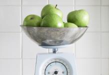 Un groupe de pommes vertes est posé dans un bol métallique sur une balance de cuisine vintage, rappelant celles que l'on trouve dans les banques d'images gratuites. La balance présente un cadran blanc avec des chiffres noirs, sur fond de murs carrelés d'un blanc immaculé.