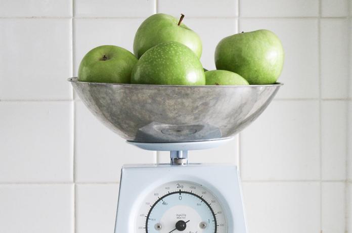 Un groupe de pommes vertes est posé dans un bol métallique sur une balance de cuisine vintage, rappelant celles que l'on trouve dans les banques d'images gratuites. La balance présente un cadran blanc avec des chiffres noirs, sur fond de murs carrelés d'un blanc immaculé.