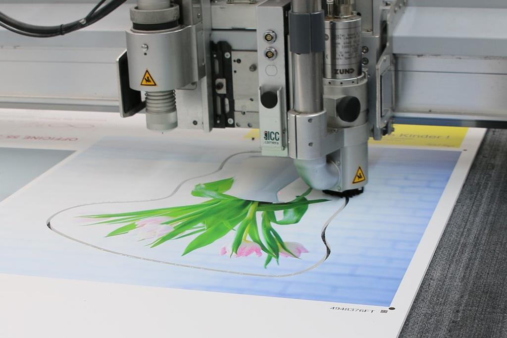 Une machine de découpe de précision découpe le contour d'un imprimé floral éclatant. L'image illustre le processus mécanique, avec des feuilles vertes et des fleurs roses méticuleusement découpées le long de leurs contours.