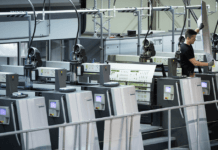 Lors d'une visite d'atelier de production offset, un ouvrier utilise une grande presse à imprimer moderne dans un environnement industriel. Plusieurs panneaux de commande sont visibles, avec des feuilles imprimées sortant de la machine. L'environnement paraît propre et organisé.