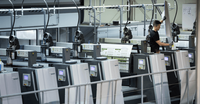 Lors d'une visite d'atelier de production offset, un ouvrier utilise une grande presse à imprimer moderne dans un environnement industriel. Plusieurs panneaux de commande sont visibles, avec des feuilles imprimées sortant de la machine. L'environnement paraît propre et organisé.