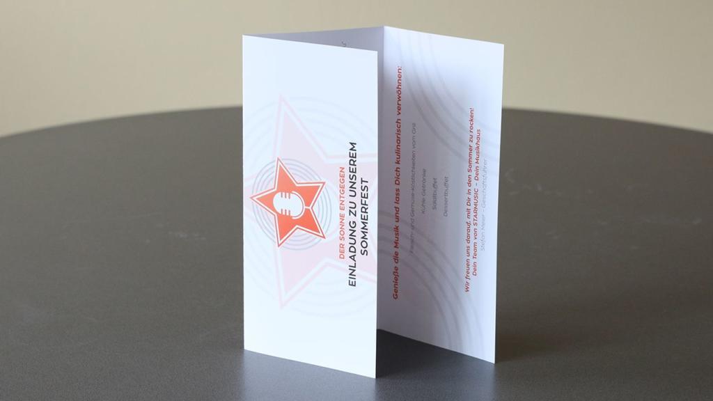Une carte d'invitation pliée, aux allures de dépliant chic, est posée sur la table. Sa surface blanche est ornée d'un microphone orange et d'un logo étoilé, avec des phrases en allemand sur un festival d'été et des jeux. Le fond est d'une douce teinte gris-jaune.