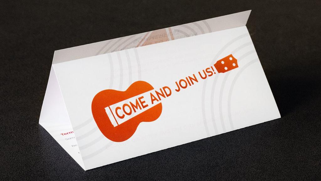 Un dépliant blanc plié sur fond noir présente une guitare orange et l'inscription en gras « COME AND JOIN US ! ». Le design est simple et accrocheur.