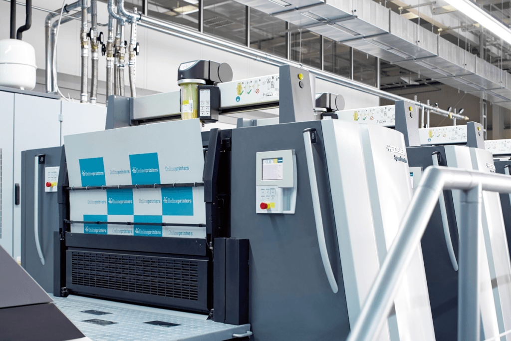 Découvrez l'atelier de production offset, une grande machine d'impression industrielle aux panneaux bleus et gris, équipée d'écrans de contrôle numériques. La machine est installée dans un environnement d'usine spacieux et lumineux, avec ses tubes métalliques et sa belle hauteur sous plafond.