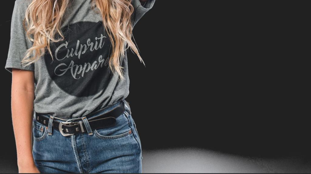 Une personne porte un t-shirt gris estampillé « Culprit Apparel », associé à un jean bleu et une ceinture noire. Le fond est noir, et l'image détourne astucieusement la chevelure, laissant la tête tronquée pour une mise en valeur élégante de la tenue.