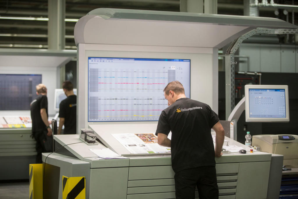 Un homme en chemise noire inspecte des feuilles imprimées sur une grande console lors d'une visite d'atelier de production offset. Deux autres ouvriers, également en chemise noire, se tiennent à l'arrière-plan au milieu d'équipements et de machines industriels.