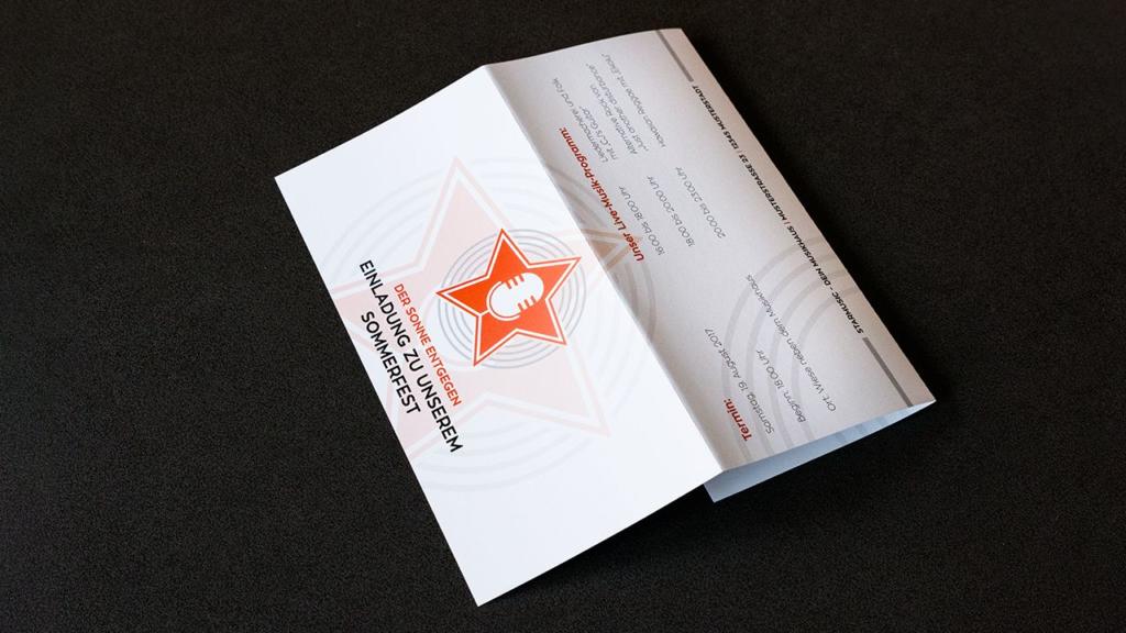 Une carte d'invitation dépliante est ouverte sur une surface sombre. La couverture présente une étoile rouge et un microphone, avec un texte en allemand. À l'intérieur, d'autres détails se dévoilent en caractères allemands. L'angle de la carte révèle à la fois la couverture saisissante et son intérieur informatif.