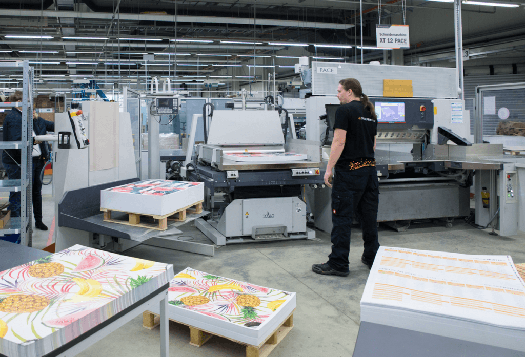 Lors d'une visite-atelier-de-production-offset, un ouvrier manipule une grande machine d'impression dans un environnement industriel. Des feuilles colorées représentant des ananas et des fleurs sont empilées sur des palettes à proximité. L'espace est bien éclairé et dispose d'équipements et de fournitures bien organisés.