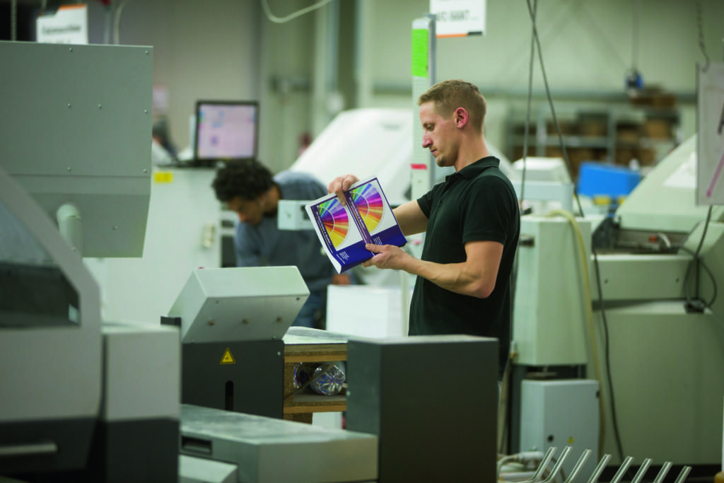 Un homme en chemise noire examine une feuille imprimée colorée dans une imprimerie animée lors d'une visite d'atelier de production offset. Il se tient parmi plusieurs machines d'impression, tandis qu'une autre personne travaille en arrière-plan. Le décor est industriel et met l'accent sur les technologies d'impression.