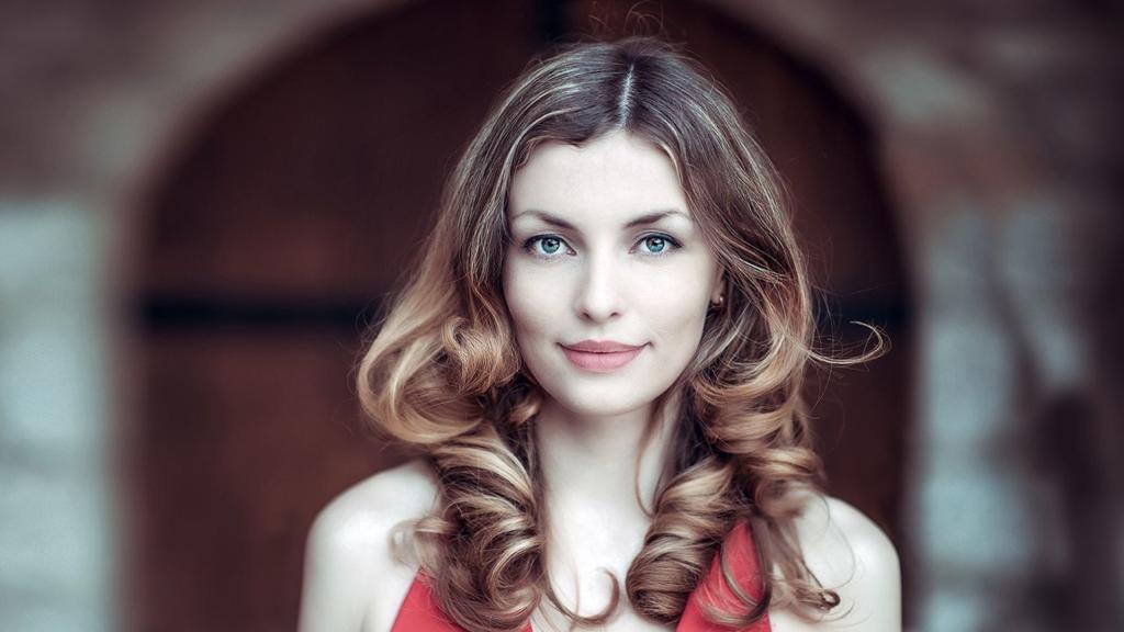 Une femme aux cheveux bruns ondulés, savamment détournés pour en rehausser l'éclat naturel, porte un haut rouge sans manches et fait face à l'objectif avec une expression neutre. Derrière elle, l'arrière-plan légèrement flou met en valeur une élégante porte en bois cintrée.
