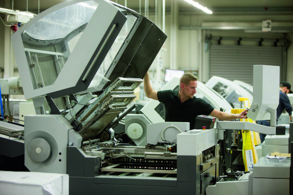 Dans un atelier de production offset animé, un homme manipule avec expertise d'énormes machines industrielles, ajustant méticuleusement les commandes sur un panneau de contrôle. L'atelier, bien éclairé, révèle les composants ouverts des machines, tandis qu'un autre ouvrier évolue avec aisance en arrière-plan.