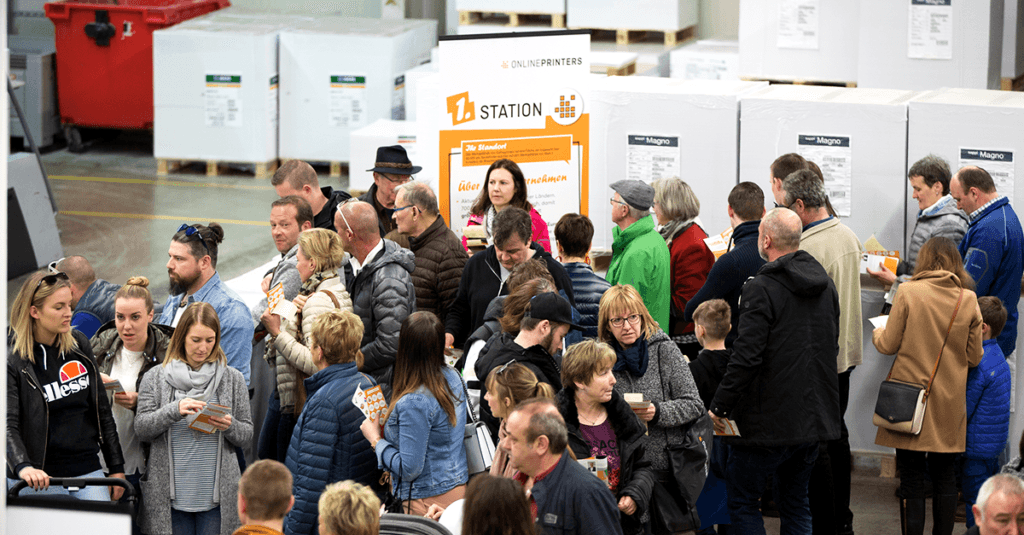 Un grand groupe de personnes faisait la queue lors d'une journée portes ouvertes près d'un panneau indiquant « Station ». Nombre d'entre eux tenaient des brochures, impatients de participer à la « visite-atelier-de-production-offset ». Le cadre dégageait une atmosphère industrielle, rappelant un entrepôt animé.