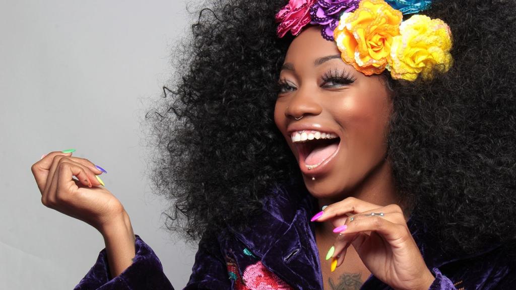 Une personne aux cheveux bouclés, ornée de fleurs colorées, arbore un sourire éclatant. Elle porte un haut en velours violet, des ongles multicolores et des piercings au visage, dégageant une énergie joyeuse et vibrante.