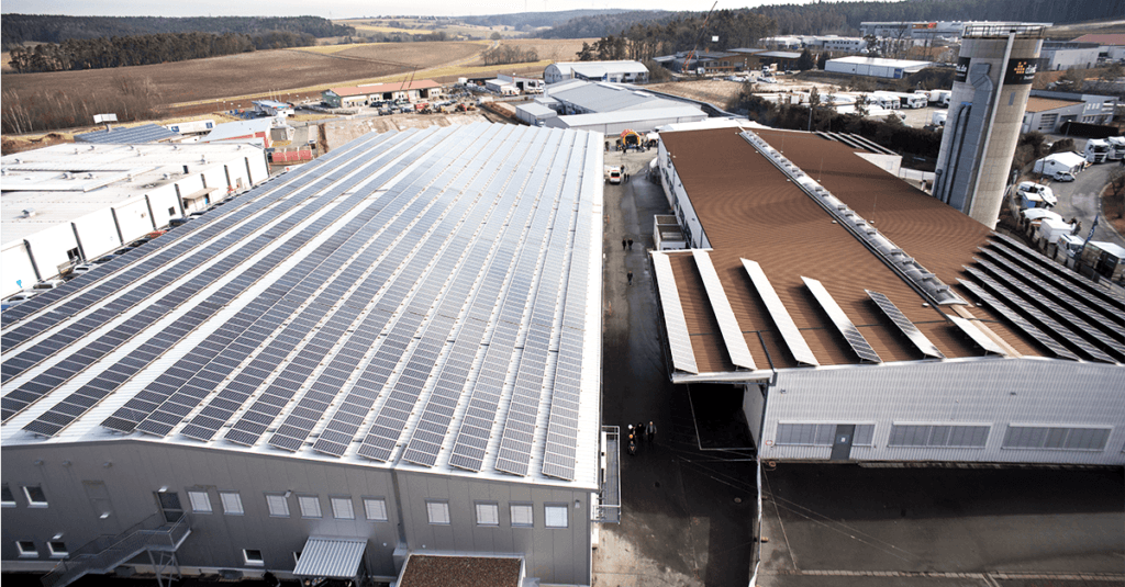 Vue aérienne de bâtiments industriels avec panneaux solaires sur le toit, comme une visite-atelier-de-production-offset. L'installation est entourée d'une zone clôturée avec des véhicules stationnés et d'un paysage forestier à proximité, soulignant ainsi l'engagement de l'entreprise en faveur des énergies renouvelables.
