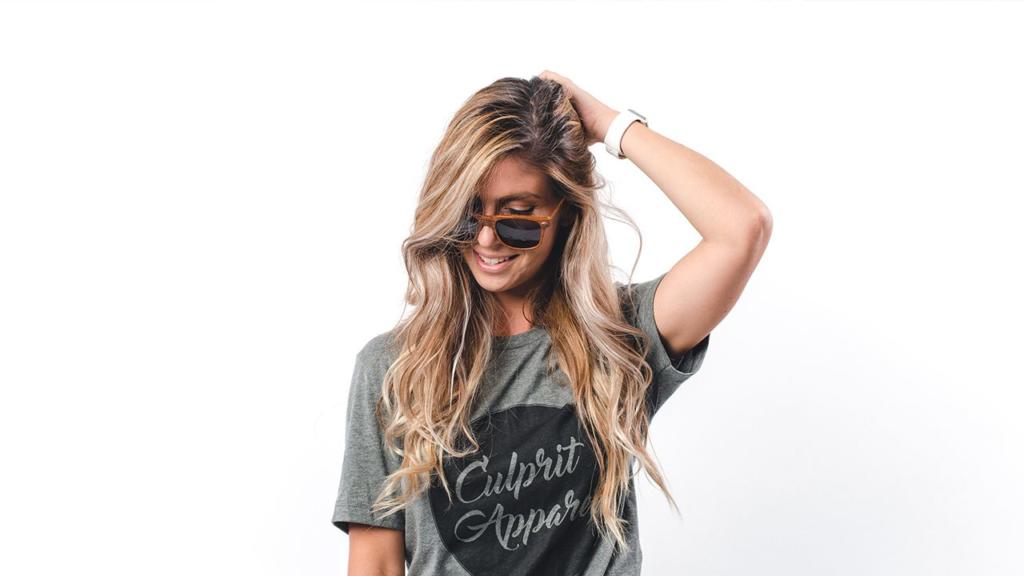Une femme aux longs cheveux blonds détournés, portant des lunettes de soleil et un t-shirt gris Culprit Apparel, sourit tandis que sa main droite touche ses cheveux. Sur fond blanc.