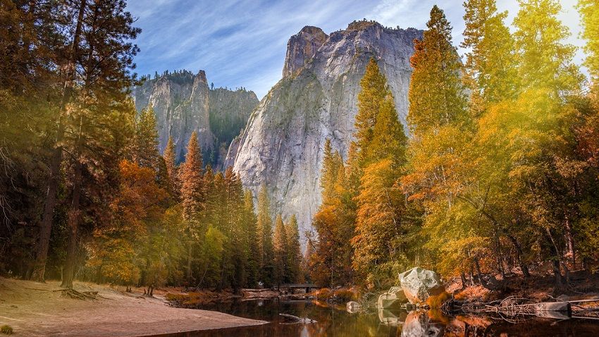 Un paysage d'automne serein peint avec Actions Photoshop met en valeur une rivière réfléchissante entourée d'arbres colorés aux teintes orange et vertes. D'imposantes falaises se dressent majestueusement en arrière-plan sous un ciel partiellement nuageux.