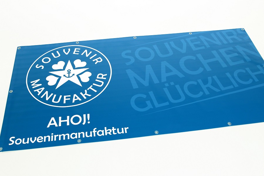 Bannière bleue avec le logo de la Manufacture Souvenir, orné d'un cœur et d'une ancre. On peut y lire « Ahoj ! Souvenirmanufaktur » et « Souvenir machen glücklich ». Le design est simple, avec du texte blanc sur fond bleu, un exemple de fabrication experte de la banderole publicitaire.