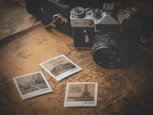 Un appareil photo Zenit-E vintage repose sur une vieille carte, entouré de trois photos imprimées : le Colisée, la Tour Eiffel et une rue animée. La dragonne renforce l'esprit de voyage nostalgique, tandis qu'une subtile vignette, réalisée avec Photoshop, apporte une touche de nostalgie intemporelle.