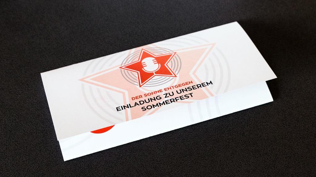Un carton d'invitation dépliant repose sur une surface sombre, affichant bien en évidence une étoile rouge avec une icône de microphone. Le texte dit : Der Sonne Entgegen Einladung zu unserem Sommerfest.