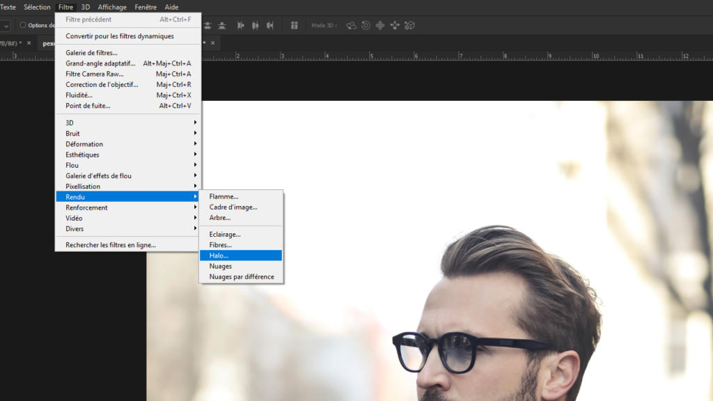 Une personne portant des lunettes et les cheveux coiffés regarde de côté, un menu logiciel se superposant à l'image. Affichant diverses options de filtres en français, comme « Rendu » et « Nuages par différence », on dirait qu'ils sont sur le point de réaliser un chef-d'œuvre avec une technique de lensflare inspirée de Photoshop.