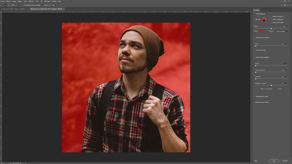 Une personne vêtue d'une chemise à carreaux rouges et d'un bonnet marron retouche sa photo sur un écran d'ordinateur, utilisant habilement Photoshop pour détourer l'image. L'interface du logiciel de retouche révèle divers outils de réglage sur la droite, tandis que l'arrière-plan de la photo arbore une texture rouge.