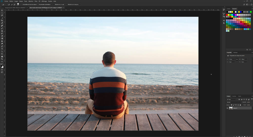 Une personne vêtue d'un pull rayé est assise sur une promenade en bois face à l'océan. Le ciel clair, avec son doux dégradé de couleurs du soleil couchant, offre un décor serein. Du sable s'étend entre la promenade et l'eau, offrant un cadre idéal pour prendre une photo avec Photoshop.