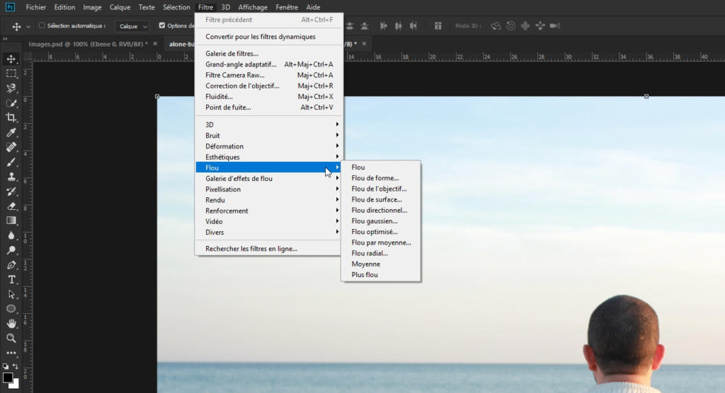 Capture d'écran d'un logiciel de retouche numérique mettant en avant le menu Filtre, présentant les options permettant de retoucher une photo avec Photoshop. Une personne portant une veste sombre se tient dans le coin inférieur droit, sur fond de ciel côtier serein et de mer calme.