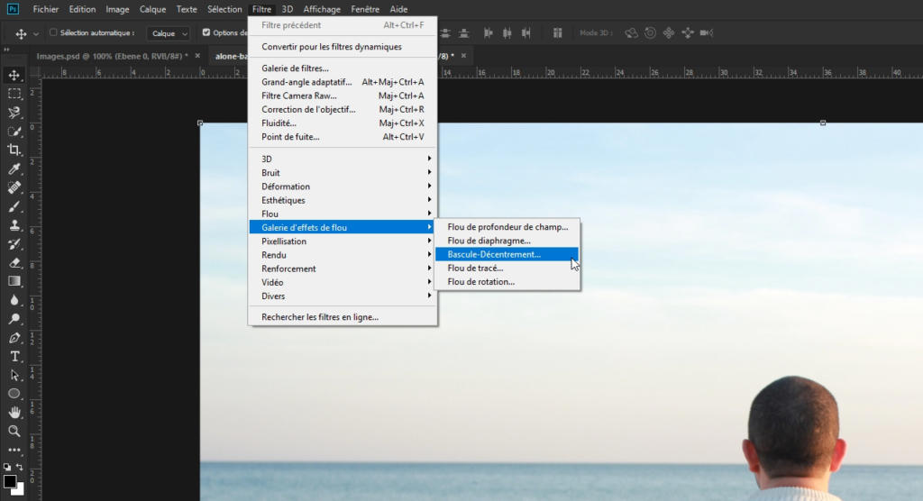 Un écran d'ordinateur présente un logiciel de retouche d'images avec un menu ouvert, révélant des options de filtres et d'effets en français. « Flouter une photo avec Photoshop » figure parmi les options. Une vue partielle d'une personne portant des lunettes apparaît en bas à droite.