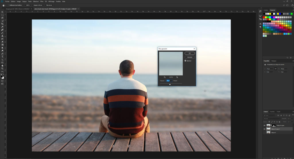 Une personne en pull rayé est assise sur une terrasse en bois face à la mer, sous un ciel dégagé. Une fenêtre d'ordinateur est superposée à l'image, montrant comment flouter une photo avec Photoshop, suggérant qu'elle est en cours de retouche avec un effet de flou.