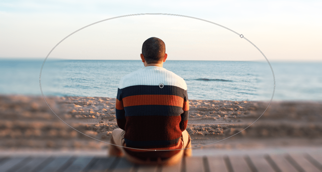 Une personne est assise sur un banc face à l'océan, vêtue d'un pull rayé multicolore. La plage et la mer calme s'étendent à perte de vue sous un ciel dégagé, créant une scène si paisible qu'on aurait envie de la capturer, et peut-être même de la retoucher subtilement avec Photoshop pour en préserver la tranquillité.