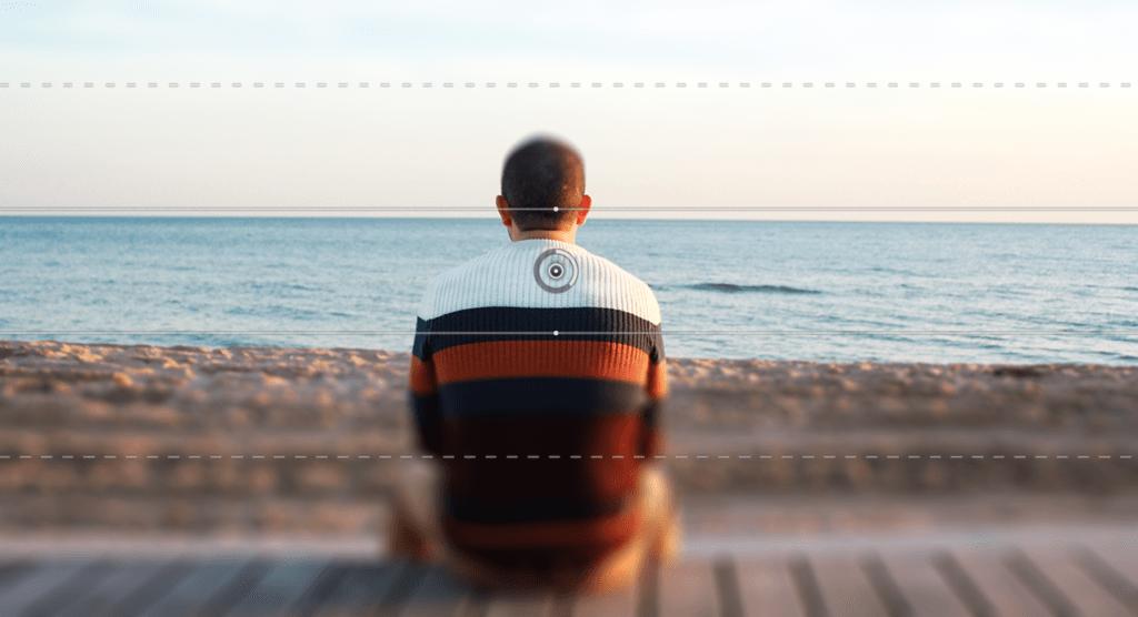 Une personne vêtue d'un pull rayé coloré est assise sur un banc en bois face à l'océan, regardant les vagues sereines et le ciel clair au coucher du soleil, envisageant d'apprendre à flouter une photo avec Photoshop pour capturer le moment de manière artistique.