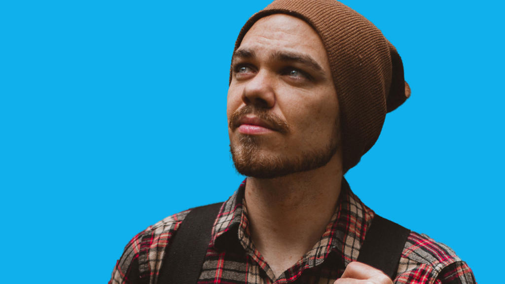 Un homme barbu, coiffé d'un bonnet marron, regarde pensivement vers le haut sur un fond bleu uni. Sa chemise à carreaux rouge et marron à bretelles accentue son air contemplatif. L'image, prête à être modifiée, constitue un excellent exemple pour ceux qui cherchent à détourer une image avec Photoshop.