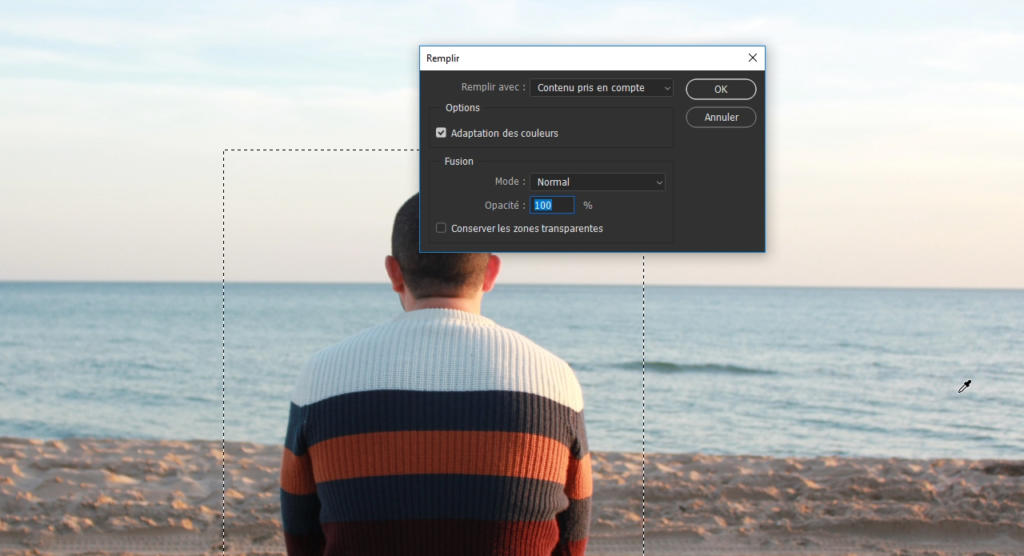 Une personne vêtue d'un pull rayé coloré est assise sur une plage tranquille, face à la mer, contemplant le menu de remplissage de Photoshop ouvert devant elle. Le ciel serein et l'eau créent un décor parfait pour apprendre à flouter une photo avec Photoshop.