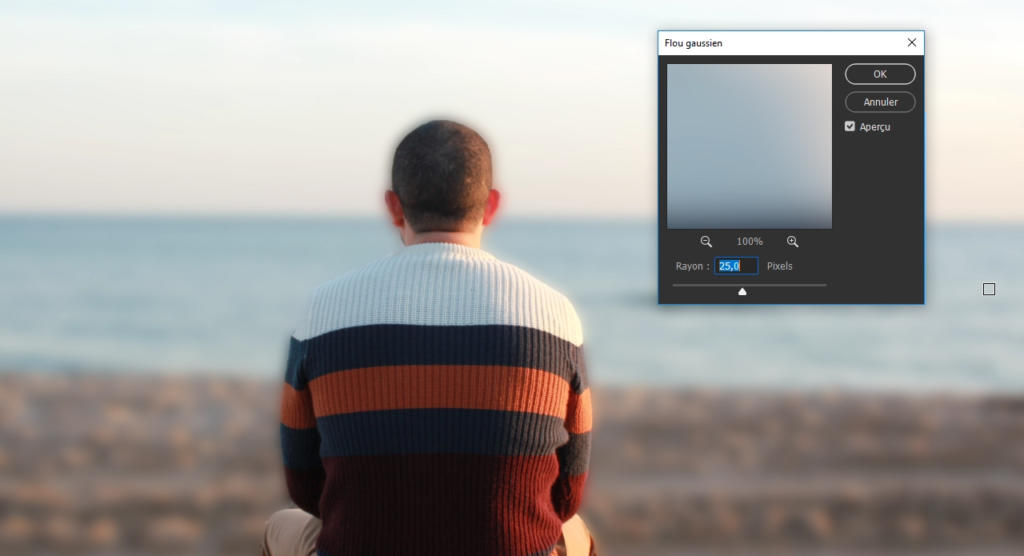 Une personne en pull rayé est assise sur le sable face à la mer, avec un effet de flou numérique rappelant flouter une photo avec Photoshop floutant partiellement la scène.