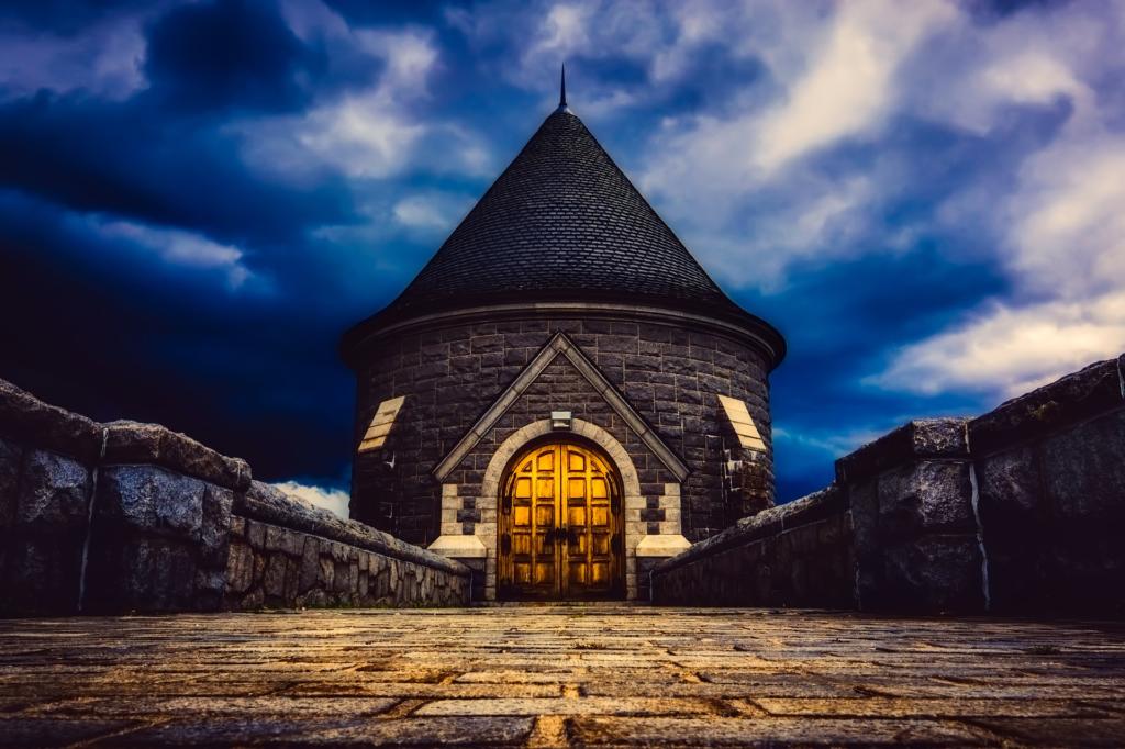 Une tour de pierre spectaculaire au toit pointu se dresse au bout d'un chemin pavé. La lueur chaleureuse de sa porte en bois contraste avec les nuages sombres et maussades en arrière-plan, comme si elle avait été retouchée par un logiciel photo pour créer une atmosphère encore plus mystérieuse.
