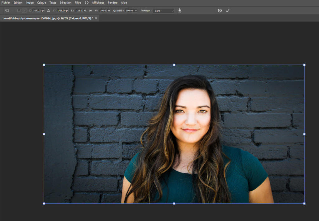 Une femme aux cheveux longs portant un haut vert se tient contre un mur de briques sombres, souriant chaleureusement à la caméra, comme si elle était prête à redimensionner l'image dans Photoshop.