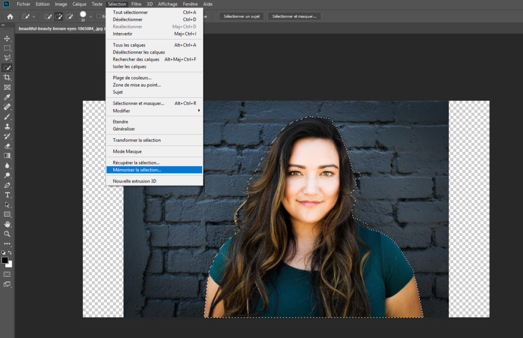 Une capture d'écran d'un logiciel de retouche photo montre une femme aux cheveux longs se tenant devant un mur de briques sombres. Sa silhouette est soulignée par une ligne pointillée, tandis qu'un menu d'options de retouche dans Photoshop est ouvert. Le curseur survole « Régler la sélection » tandis que l'on examine l'option permettant de redimensionner l'image.