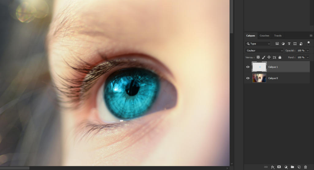Gros plan sur un œil bleu vif avec un flou artistique sur les cils. L'image est affichée sur un écran de retouche numérique, affichant les calques et les options de retouche à droite. L'œil et la peau environnante sont mis en valeur par un éclairage naturel.
