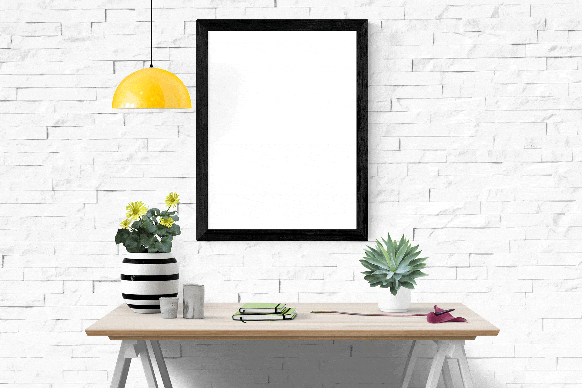 Espace de travail minimaliste avec une table en bois, des plantes en pot, un carnet fermé avec des lunettes et un petit marteau. Un cadre vide est accroché à un mur de briques blanches. Une suspension jaune, au-dessus, apporte une touche de couleur.