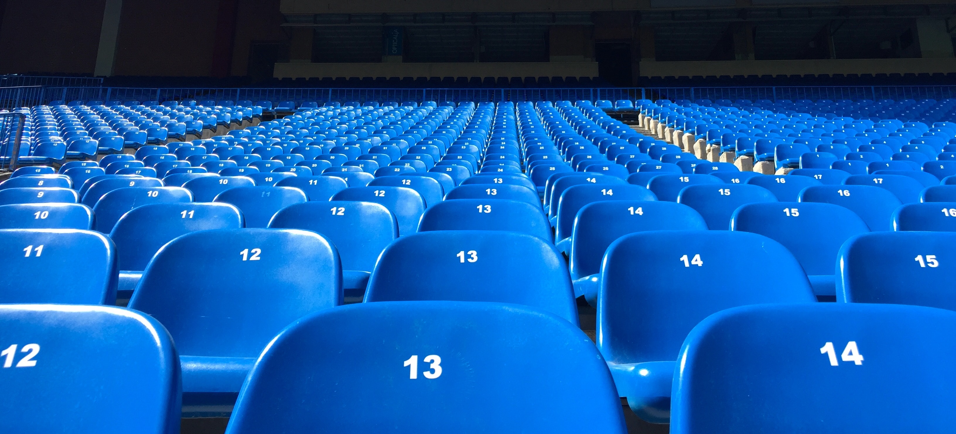 Des rangées de sièges bleus vides sont soigneusement disposées, avec des numéros comme 12, 13 et 14 bien visibles. La lumière vive du soleil illumine les sièges et projette des ombres. Ce cadre serein laisse présager l'excitation à venir, les supporters attendant avec impatience les événements décrits dans le plan de jeu de l'EM 2021.