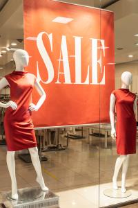 Deux mannequins en robes rouges se tiennent devant une grande pancarte rouge « SOLDES » dans la vitrine d'un magasin. L'intérieur du magasin est visible en arrière-plan, avec ses portants et son éclairage intense.