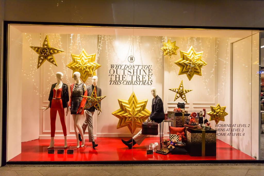 Vitrine avec mannequins en tenues festives. Des étoiles dorées les entourent. Au mur, on peut lire : « Pourquoi ne pas faire plus de lumière que le sapin à Noël ? » Des cartons sont empilés aux pieds des mannequins, sur un sol rouge.