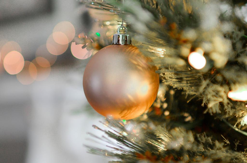 Gros plan sur une branche de sapin de Noël décorée d'une douce décoration rose. Des lumières chaudes et floues scintillent doucement en arrière-plan, créant une atmosphère chaleureuse et festive.