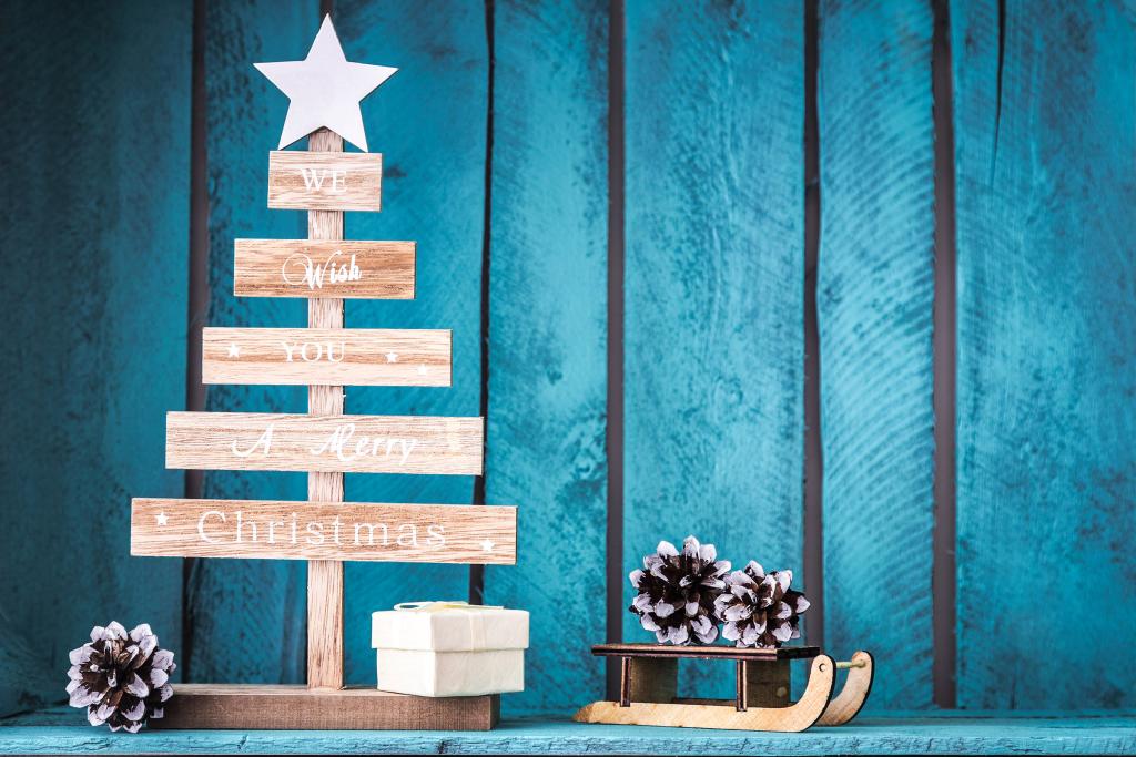 Une décoration de sapin de Noël en bois, surmontée d'une étoile, se détache sur un fond de bois bleu. Elle porte le message « Nous vous souhaitons un joyeux Noël ». À proximité se trouvent une boîte cadeau, une petite luge et des pommes de pin.