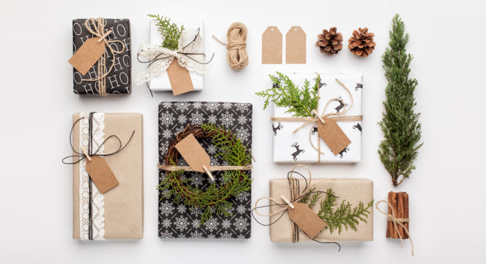 Une collection de cadeaux soigneusement emballés avec du papier kraft et des nœuds en ficelle, décorés de pommes de pin, de brindilles de verdure et de petites couronnes. Des étiquettes et de la ficelle sont disposées à côté des cadeaux sur un fond blanc.
