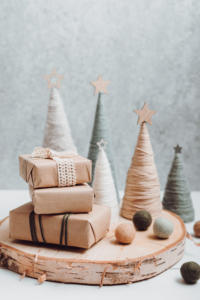 Une scène de Noël chaleureuse avec de petits sapins recouverts de laine et surmontés d'étoiles, et trois cadeaux emballés posés sur une surface en bois. Des boules de laine sont éparpillées un peu partout, créant une atmosphère minimaliste et festive.
