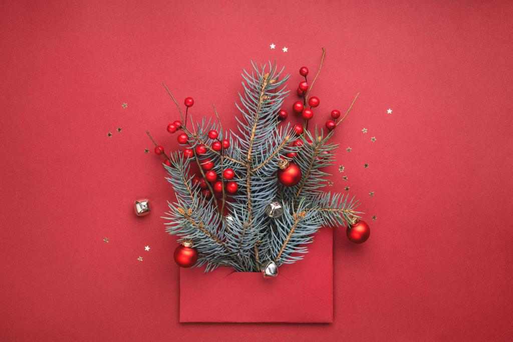 Enveloppe de Noël rouge ornée de branches de sapin, de baies rouges et de boules métalliques sur fond rouge. Étoiles argentées et grelots sont disséminés un peu partout, créant une ambiance festive.
