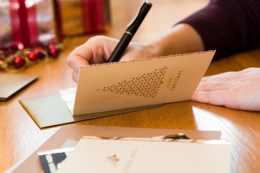 Une personne écrit sur une carte de Noël au stylo. La carte représente un sapin de Noël stylisé composé de petites formes découpées et le texte « Joyeux Noël ». Des cadeaux emballés dans du papier festif et des rubans apparaissent flous en arrière-plan.