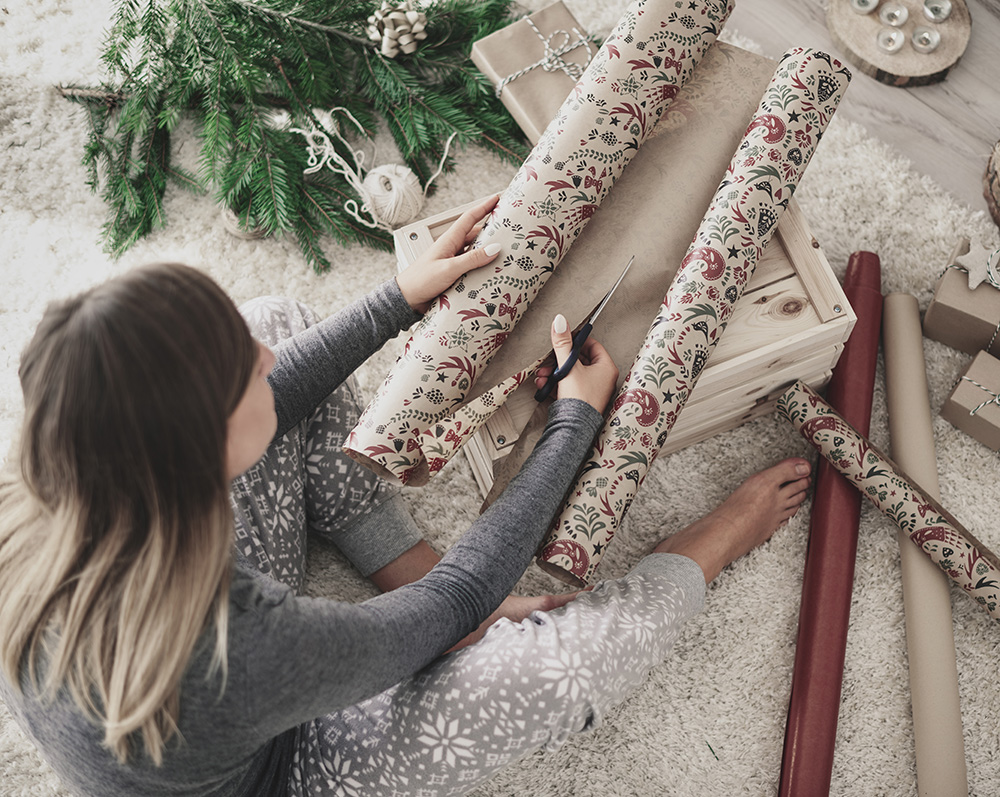 Assise sur un tapis, une personne en pyjama confortable découpe du papier cadeau décoratif avec des ciseaux. Des branches de pin et des décorations sont disposées à proximité, et quelques boîtes emballées sont disposées autour. La scène évoque une ambiance festive et chaleureuse.