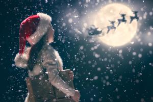 Une personne portant un bonnet de Père Noël et tenant un coffret cadeau contemple la pleine lune. Devant elle, les silhouettes d'un traîneau et de rennes volent dans un ciel nocturne enneigé.